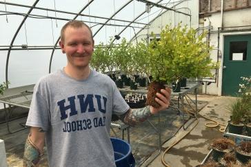 Male student in greenhouse holds a plant. His t-shirt says UNH Graduate School.